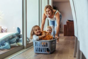 Mother playfully pushing her children in a laundry basket across the floor.
