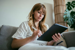 Woman using a stylus writing on a digital tablet while sitting on her couch.